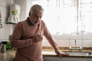 A senior man holds a glass of water in the kitchen and wonders about the link between depression and heart disease