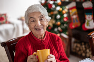 Knowing the signs to monitor for wellbeing and safety in older loved ones helps them to continue to enjoy the comfort of home, as this older woman is doing while enjoying a cup of coffee in front of her holiday decorations.